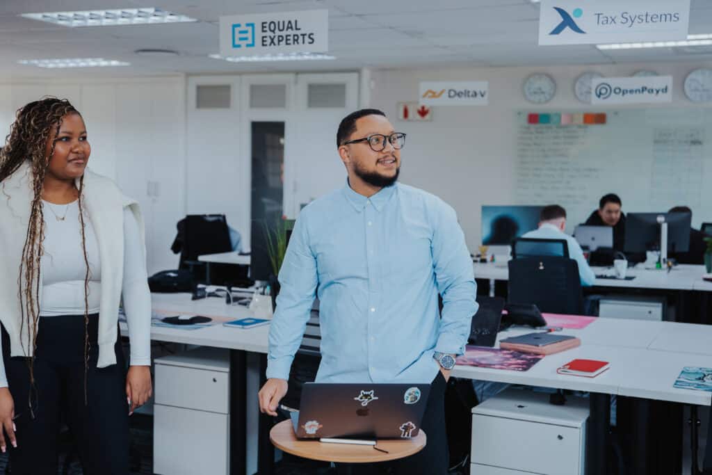 Two Delta v team members in conversation in the company's open-plan office, with the Delta v logo and a busy sales floor visible behind them.